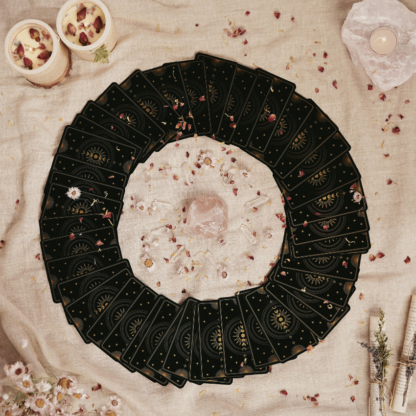 A circle of Tarot cards sits on a tablecloth with flower petals and crystals scattered around it.