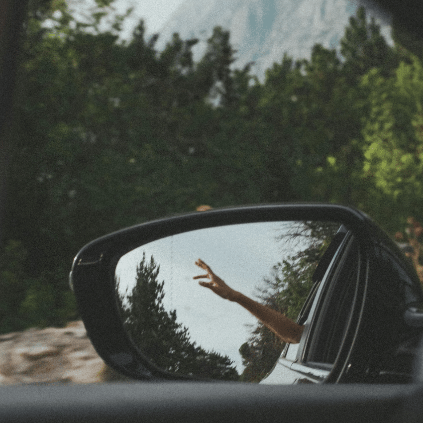 A hand point towards the sky in a rear view mirror of the car. past the mirror is a forest and mountain to represent the final week of Taurus season energy for this week