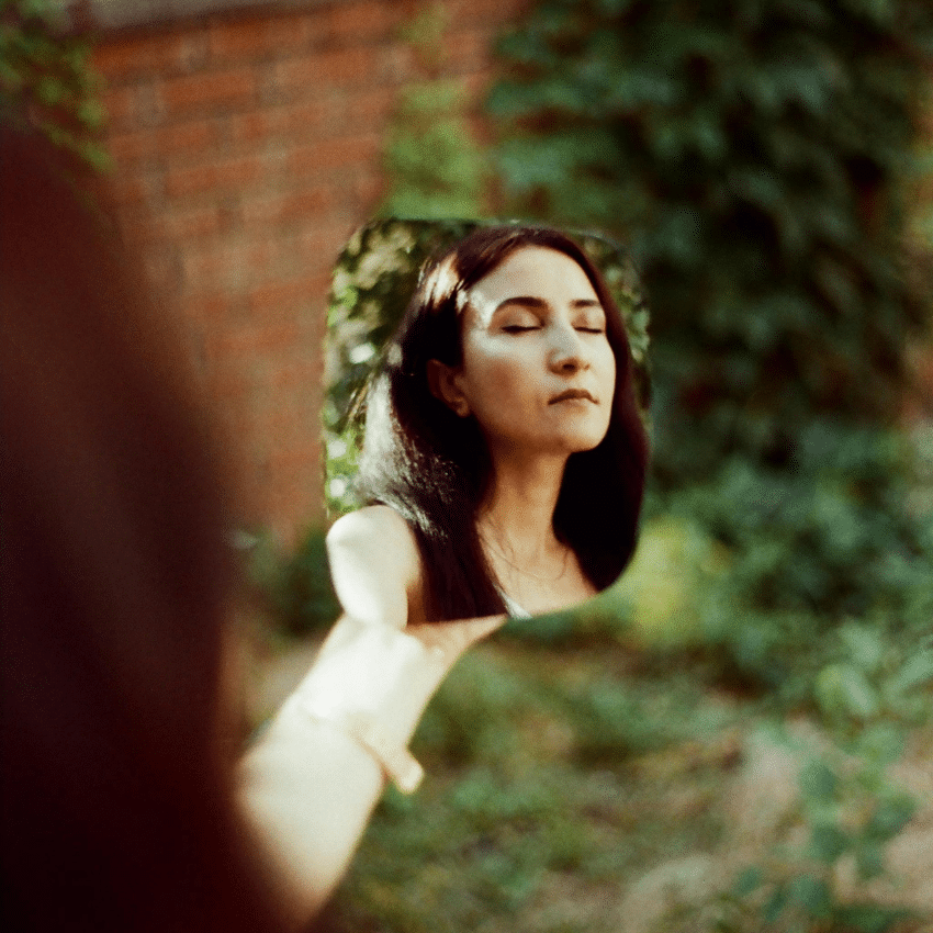 A brunette woman looks in a mirror, her reflection shown to the viewer. Her eyes are closed as if in deep thinking.