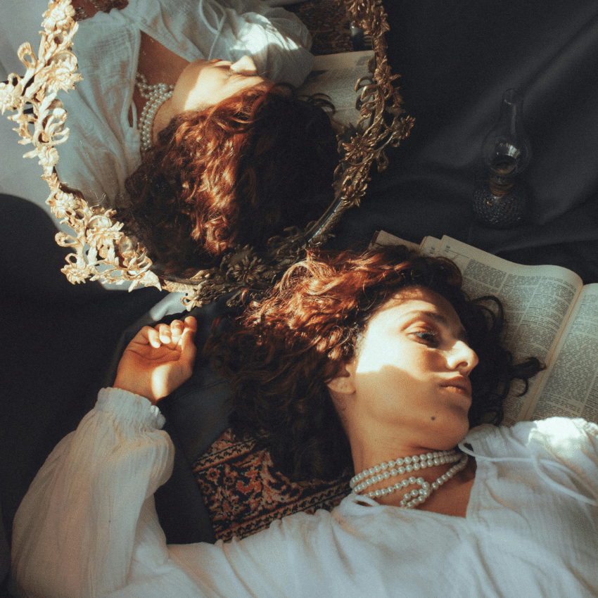 A woman wearing a white shirt and pearls lays in front of a mirror, her reflection lit up in the Sun.