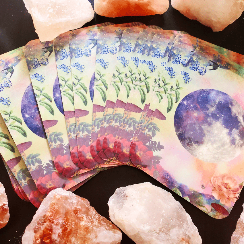 A colorful Tarot deck is spread out on a black table with pink crystals surrounding them.