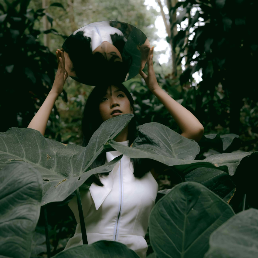 A woman stands amongst large green leaves holding a mirror above her head to represent that reflections to be done using this week