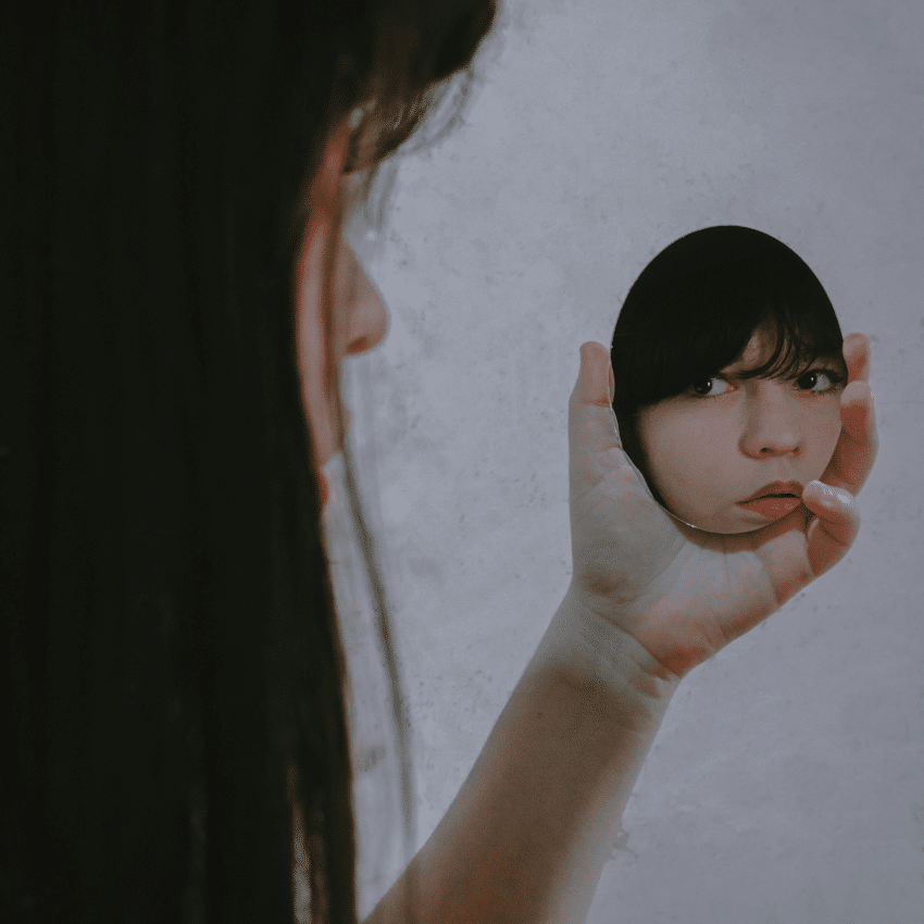 A woman looks into a small handheld mirror, her reflection looking back at the viewer to represent the reflection to be done with this week