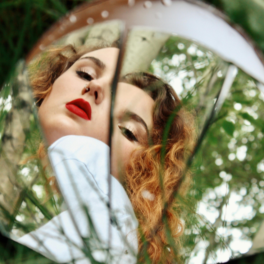 A woman with red hair and red lipstick looks in a circular mirror, her reflection representing the weekly astrology energy.
