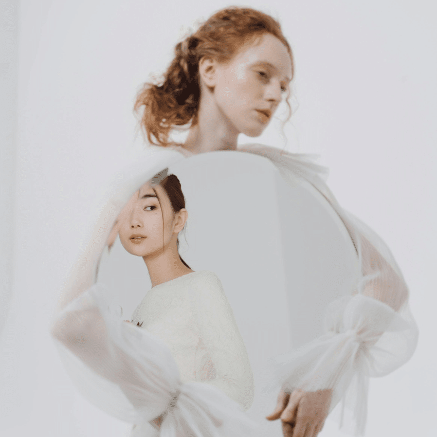 A woman with red hair holds a mirror, reflecting an Asian woman with dark hair. This represents the shift from Capricorn to Aquarius energy during this week