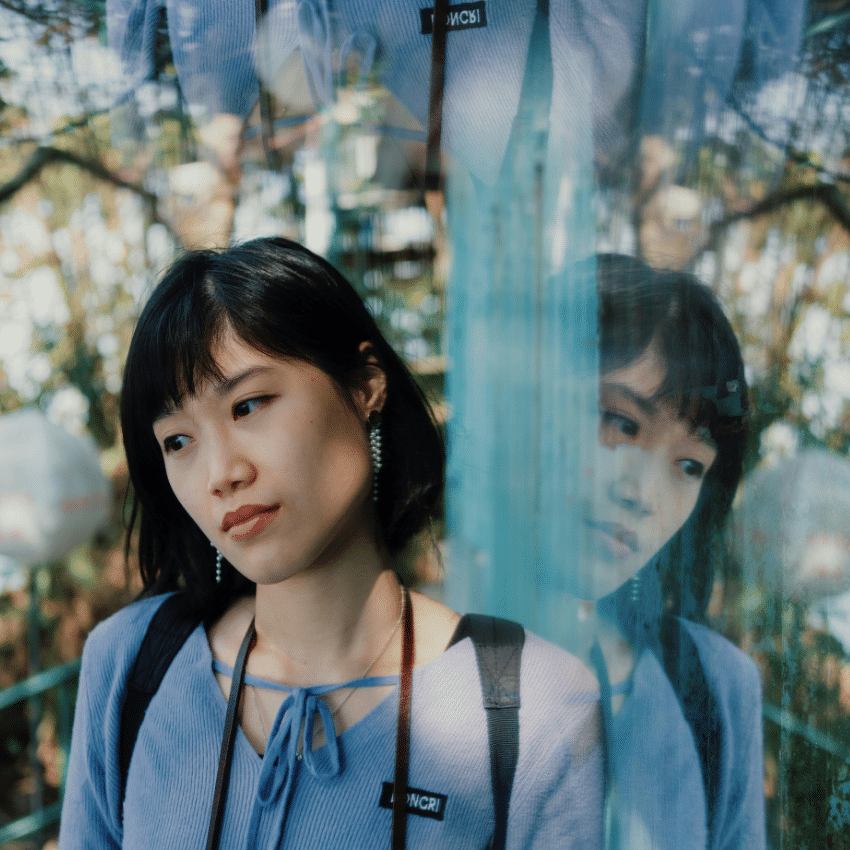 a woman looks off to her right side, her reflection showing in a mirrow or panel of glass beside her.