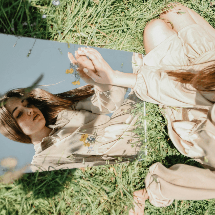 A woman with long brown hair sits on grass and in a looks at a mirror , her downcast reflection representing this weeks astrology reflections.