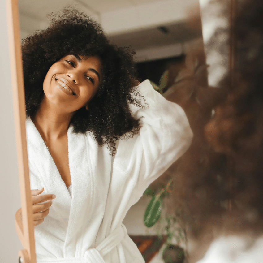 A black woman with a curly afro wearing a white robe looks into a mirror, her smiling reflection representing this week