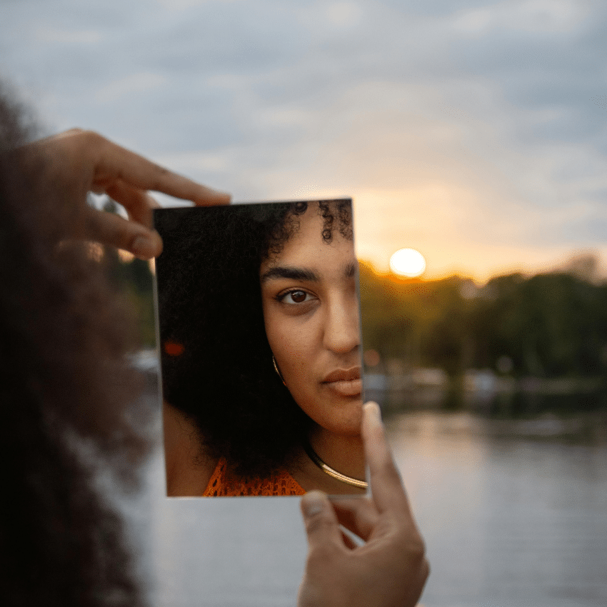 A black woman looks in a handheld mirror, her reflection appearing in front of a body of water and a setting sun, representing this week