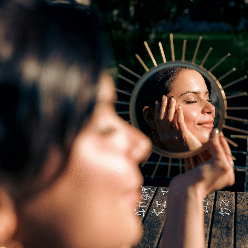 A woman faces away from a mirror, her reflection showing her cupping the side of her face.