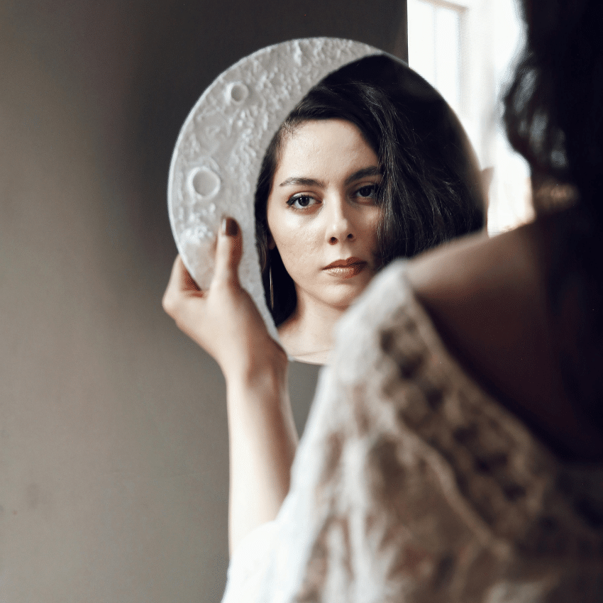 A woman looks in a mirror framed by a Moon decor shape representing the energy of this week's astrology.