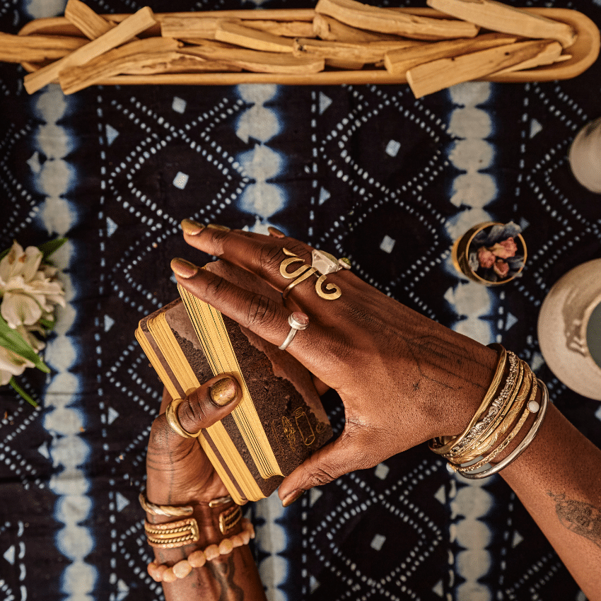 A woman's hands hold a deck of Tarot cards, poised to shuffle them. She is braced above a mystical looking table cloth, crystals, and wood for burning to represent the energy of June.