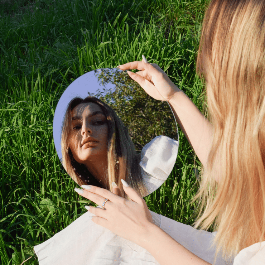 A woman with blonde hair looks in a mirror on a patch of green grass.